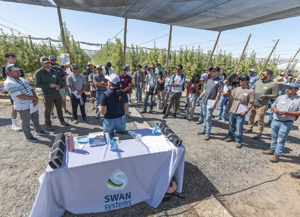 En julio, Alejandra Odegard, de Swan Systems, habla sobre riego automatizado con los asistentes al día de campo del proyecto Smart Orchard de la Universidad Estatal de Washington en Mattawa. Los investigadores afirman que las herramientas de Swan y otras empresas les permitieron regar y nebulizar una parcela de manzanos de forma automática con menos agua y obtener resultados de producción similares, o incluso mejores, que en una parcela controlada por el productor. (TJ Mullinax/Good Fruit Grower)