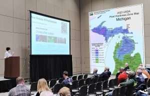 Michigan State University tree fruit integrator Jackie Perkins discusses changing weather and pest patterns during the Great Lakes EXPO in December. Here, she was describing a new MSU website called mifruitdecision.com, which helps growers manage climate-related pest risks. On the right is an illustration of another one of her presentation slides, the 2023 USDA Plant Hardiness Zone Map of Michigan, which she used to illustrate changing climate patterns. (Photo illustration by Matt Milkovich and TJ Mullinax/Good Fruit Grower)