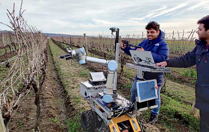 Carnegie Mellon University graduate students Nomaan, left, and Sparsh Garg operate a pruning robot at a Cornell University research vineyard in Portland, New York. Nomaan is teleoperating the small robotic arm that is guiding the movements of the large robotic pruning arm. The researchers are gathering demonstration data to train robots to prune vines more like humans do. (Courtesy Carnegie Mellon University)